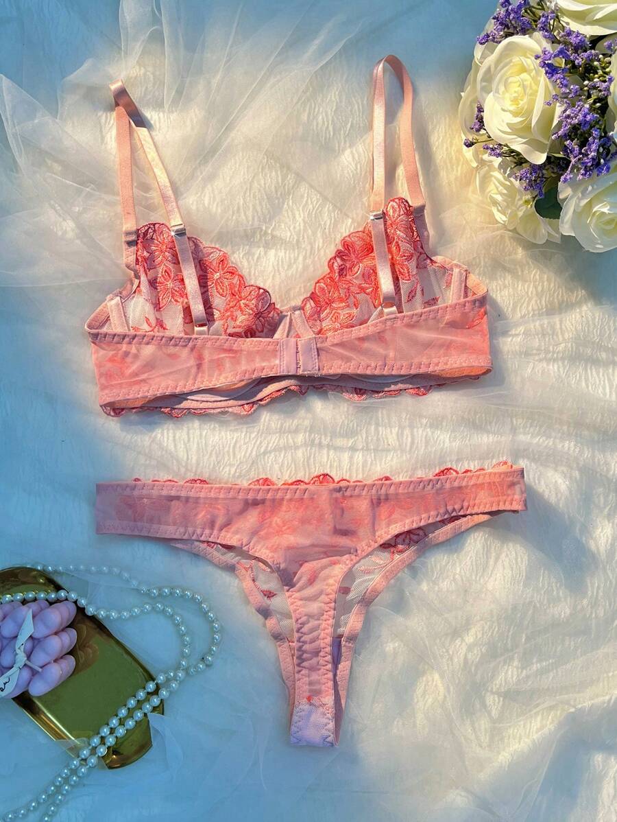 Conjunto de lencería romántico con bordado floral rojo sobre malla rosa, exhibido en un gancho decorativo.