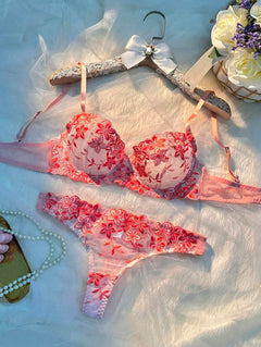 Conjunto de lencería romántico con bordado floral rojo sobre malla rosa, exhibido en un gancho decorativo.