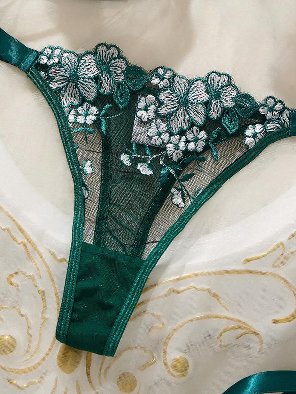 Vista frontal de la tanga de lencería verde con malla transparente, bordado floral y cintas de satén a los lados.
