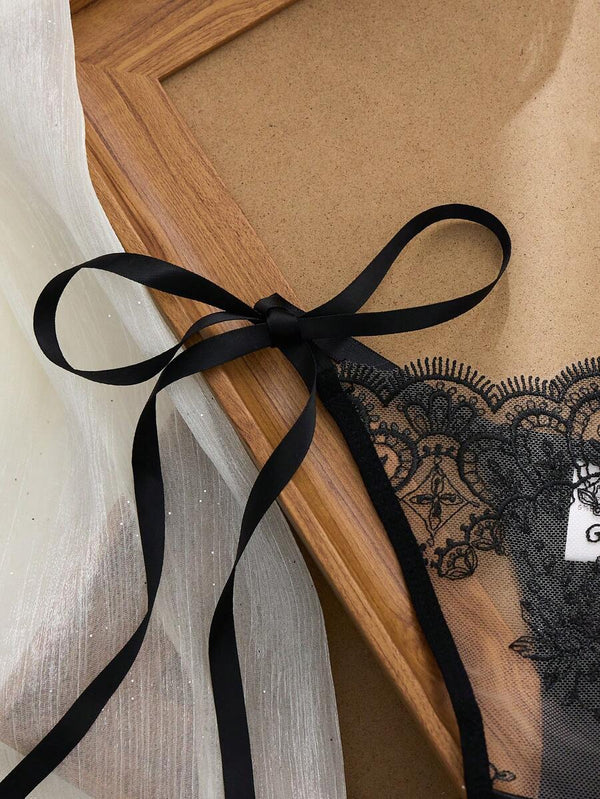 Detalle del panti tipo tanga de encaje negro con lazos de satín ajustables a los lados sobre una tela blanca.