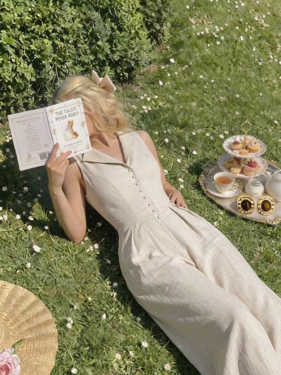 Mujer vistiendo un jumpsuit beige sin mangas de pierna ancha, recostada en el césped leyendo un libro.