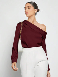 Mujer modelando una blusa elegante color burdeos con cuello asimétrico y hombro descubierto, combinada con pantalones blancos.