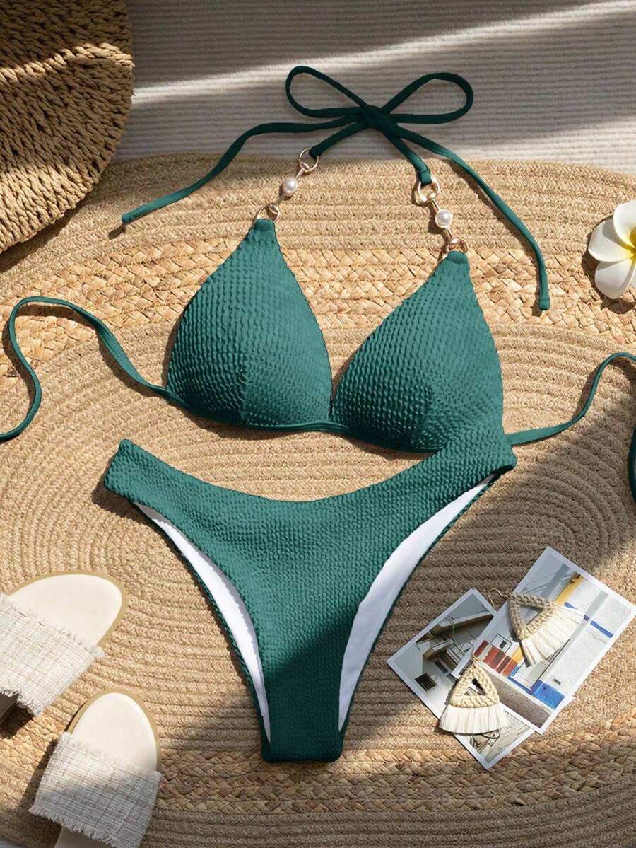 Bikini de dos piezas color verde oscuro con textura y detalle de perlas en los tirantes, visto desde arriba sobre una estera de paja.