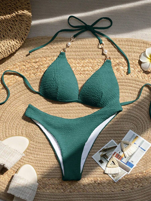 Bikini de dos piezas color verde oscuro con textura y detalle de perlas en los tirantes, visto desde arriba sobre una estera de paja.