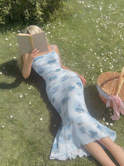 Mujer rubia recostada en el césped leyendo un libro mientras luce un vestido midi celeste con estampado floral azul.