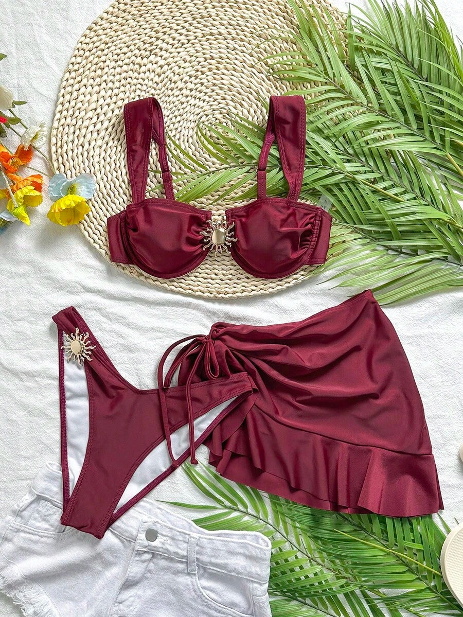 Set de traje de baño de tres piezas color vino tinto sobre una superficie blanca y una base de paja.