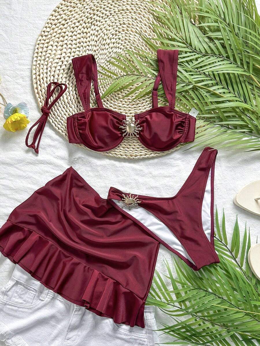 Set de traje de baño de tres piezas color vino tinto sobre una superficie blanca y una base de paja.
