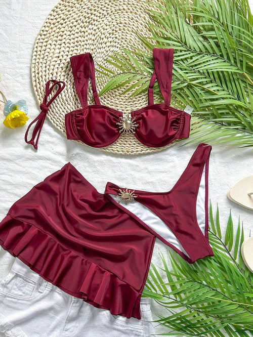Set de traje de baño de tres piezas color vino tinto sobre una superficie blanca y una base de paja.