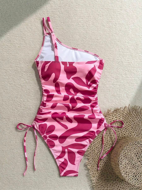 Primer plano del tirante asimétrico y el estampado vibrante del traje de baño rosa junto a unas flores y gafas de sol.