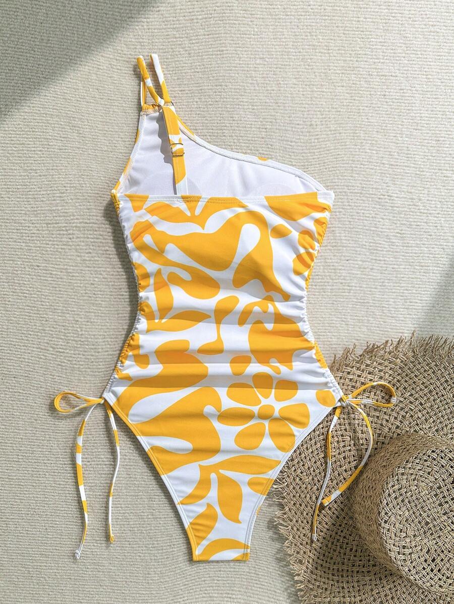 Bañador asimétrico de una pieza en color blanco y amarillo con estampado floral abstracto sobre arena y accesorios.