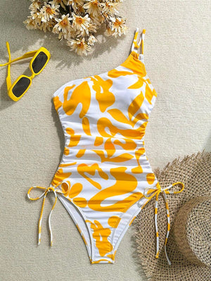 Bañador asimétrico de una pieza en color blanco y amarillo con estampado floral abstracto sobre arena y accesorios.