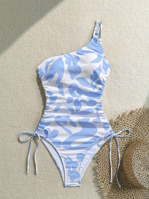 Traje de baño asimétrico con estampado abstracto junto a un sombrero de paja y gafas de sol amarillas, creando un look de playa.