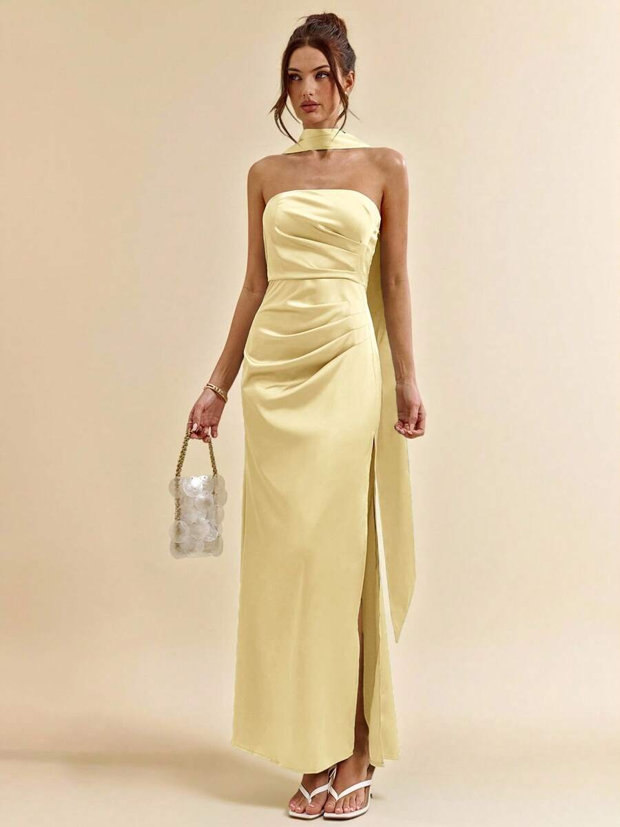 Mujer posando con un vestido largo satinado amarillo pastel de cuello halter y abertura lateral.