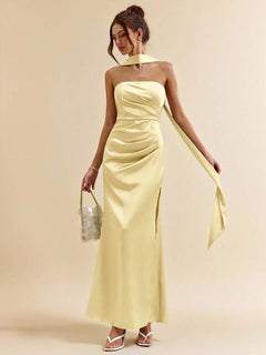 Mujer posando con un vestido largo satinado amarillo pastel de cuello halter y abertura lateral.
