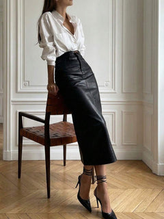 Mujer con camisa blanca y falda midi negra de piel sintética posando elegantemente apoyada en una silla de madera.