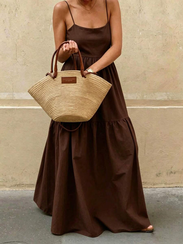 Mujer caminando con un vestido largo de verano color café mostrando el movimiento de la falda y un bolso de mano de fibra natural.