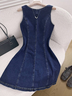 Vestido de mezclilla azul oscuro sin mangas con corte A, visto desde el frente sobre un sillón texturizado.