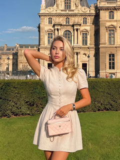 Mujer rubia con vestido corto beige de manga corta y cuello alto posando al aire libre.