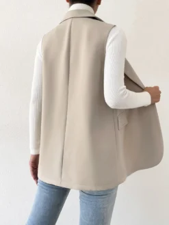 Chaleco largo beige con botones dorados y solapas clásicas, combinado con un suéter de cuello alto y jeans.